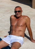 Man sitting by a pool wearing sunglasses and white shorts on a sunny day.